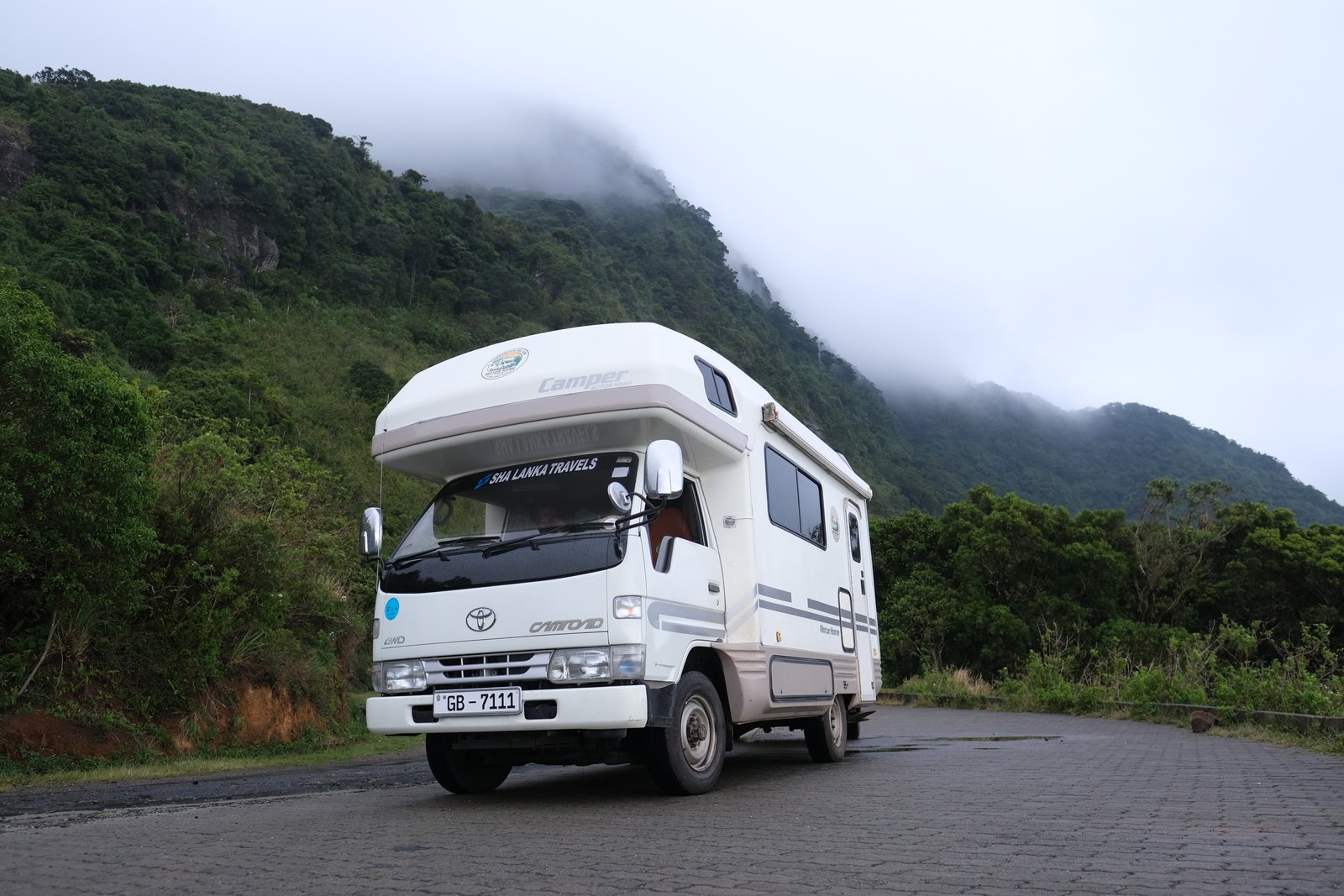 The four-wheel-drive camper on a mountain road with mist rolling over the forested slopes behind it