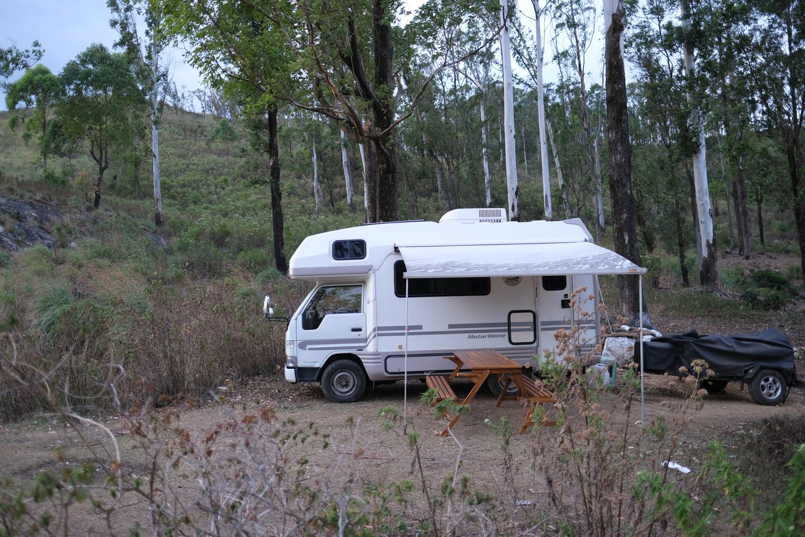 Camper parked among eucalyptus trees with its awning out and a wooden picnic table set up beside it