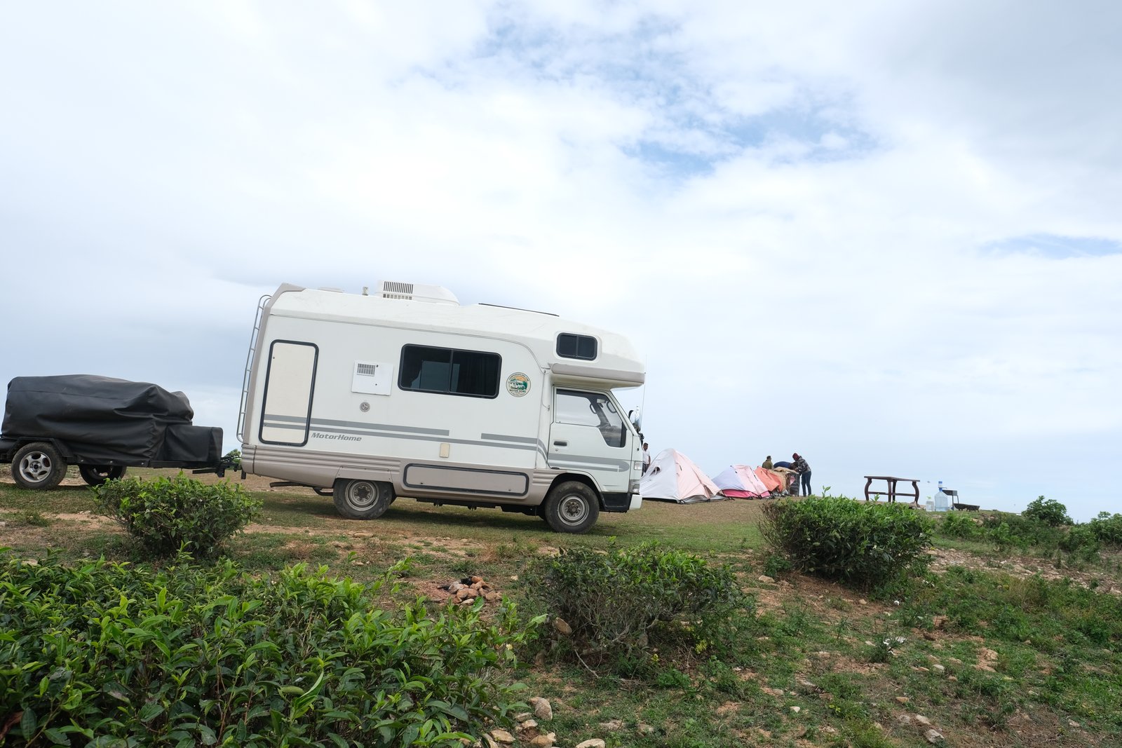 Camper and tents pitched on a grassy hilltop among tea bushes as travellers set up camp